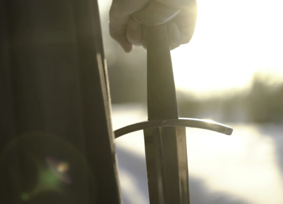 Man in medieval suit holding sword. Action. Close-up of man's hand in medieval suit holding sword hilt on background of sunlight. Epic view of medieval hero