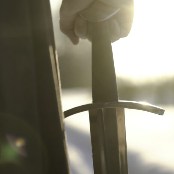 Man in medieval suit holding sword. Action. Close-up of man's hand in medieval suit holding sword hilt on background of sunlight. Epic view of medieval hero