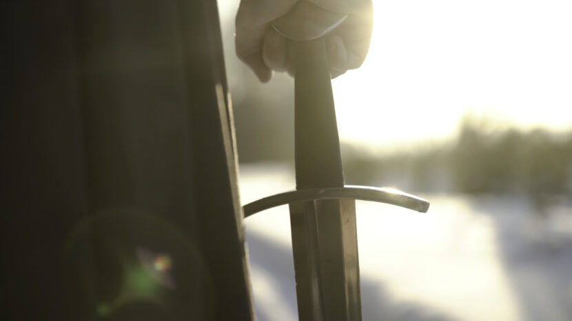Man in medieval suit holding sword. Action. Close-up of man's hand in medieval suit holding sword hilt on background of sunlight. Epic view of medieval hero